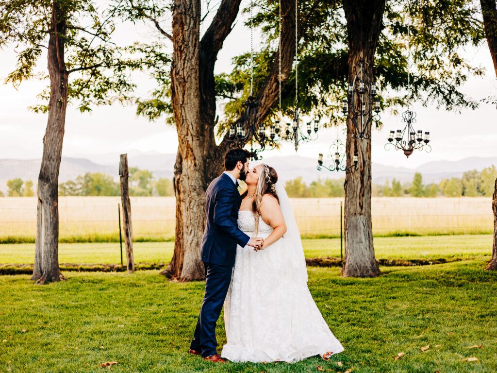 A photo of a groom wearing a blue suit and a bride wearing a strapless white wedding dress kiss under the chandeliers at Shupe Homestead in Longmont, CO. 