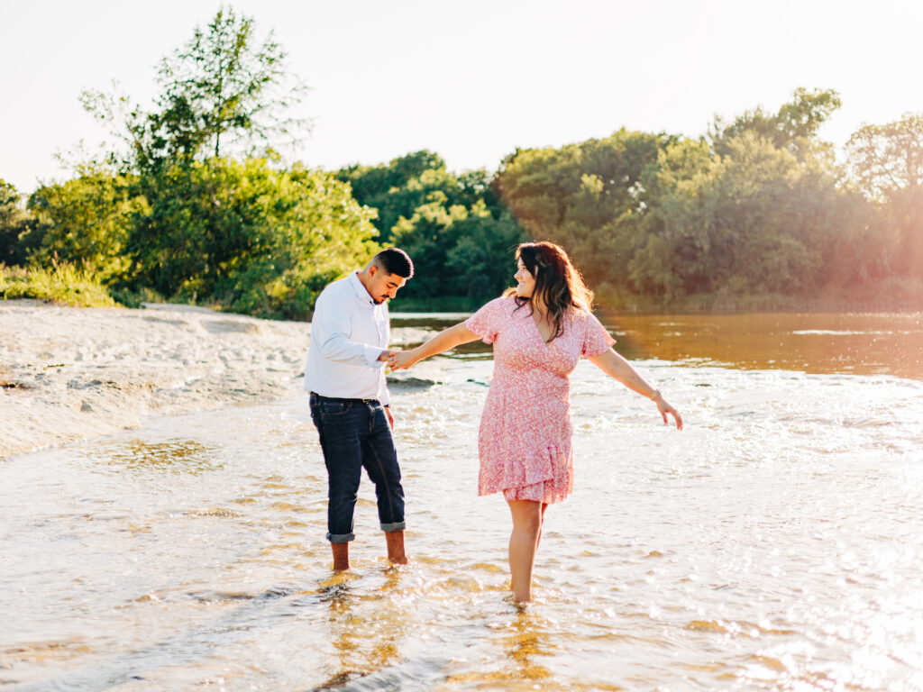 A couple wading through water during an engagement session during the summer. The woman is wearing a pink patterned dress and the man is wearing jeans that are rolled up and a button-up shirt.