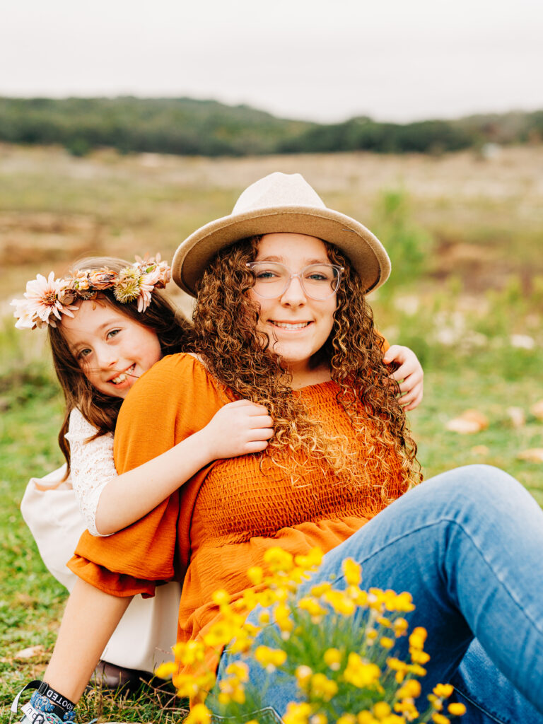 Two sisters at a fall family session at Pace Bend Park in Austin, TX. The youngest sister wearing a flower crown wraps around the oldest sister from behind.