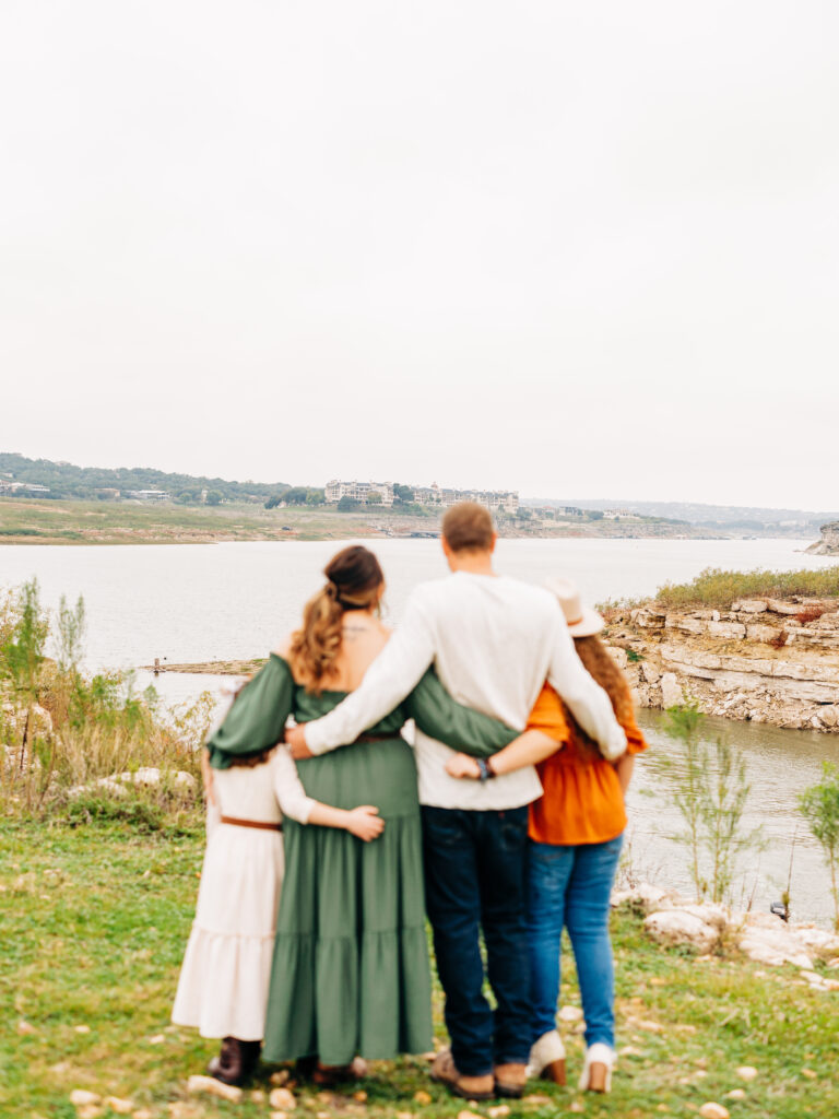A family of four in Autumn at Pace Bend Park in Austin, TX. Lake Travis is the subject of the image as the family hugs eachother.