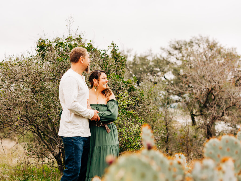 A couple looks off into the distance during a family session at Pace Bend Park in Austin, TX. There is a cactus visible in the foreground of the image.