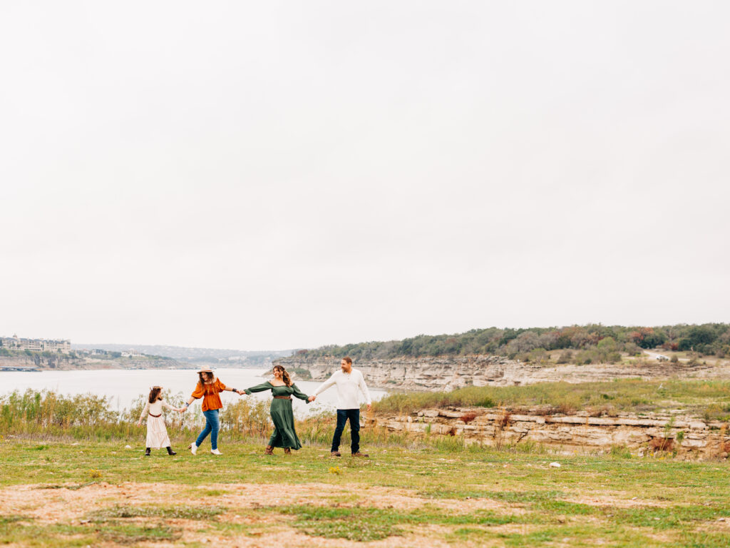 A family of four in Autumn at Pace Bend Park in Austin, TX. Lake Travis is visible in the background as the family walks, holding hands.