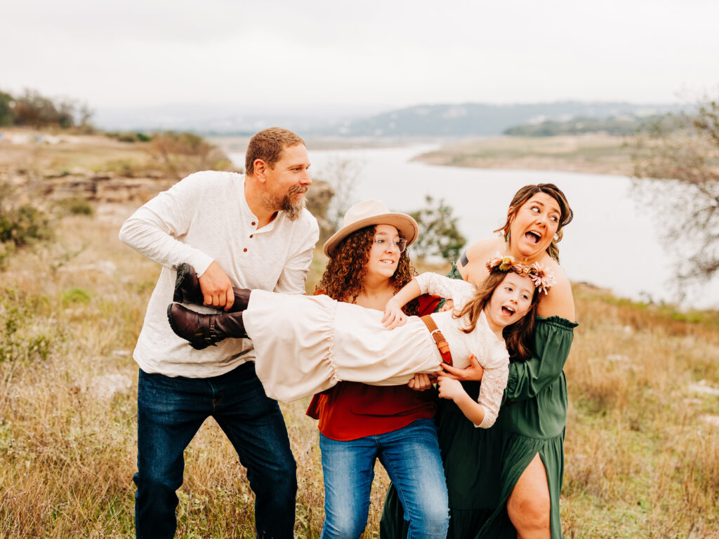 A family of four in Autumn at Pace Bend Park in Austin, TX. Lake Travis is visible in the background as the three oldest hold the youngest, pretending to throw her.