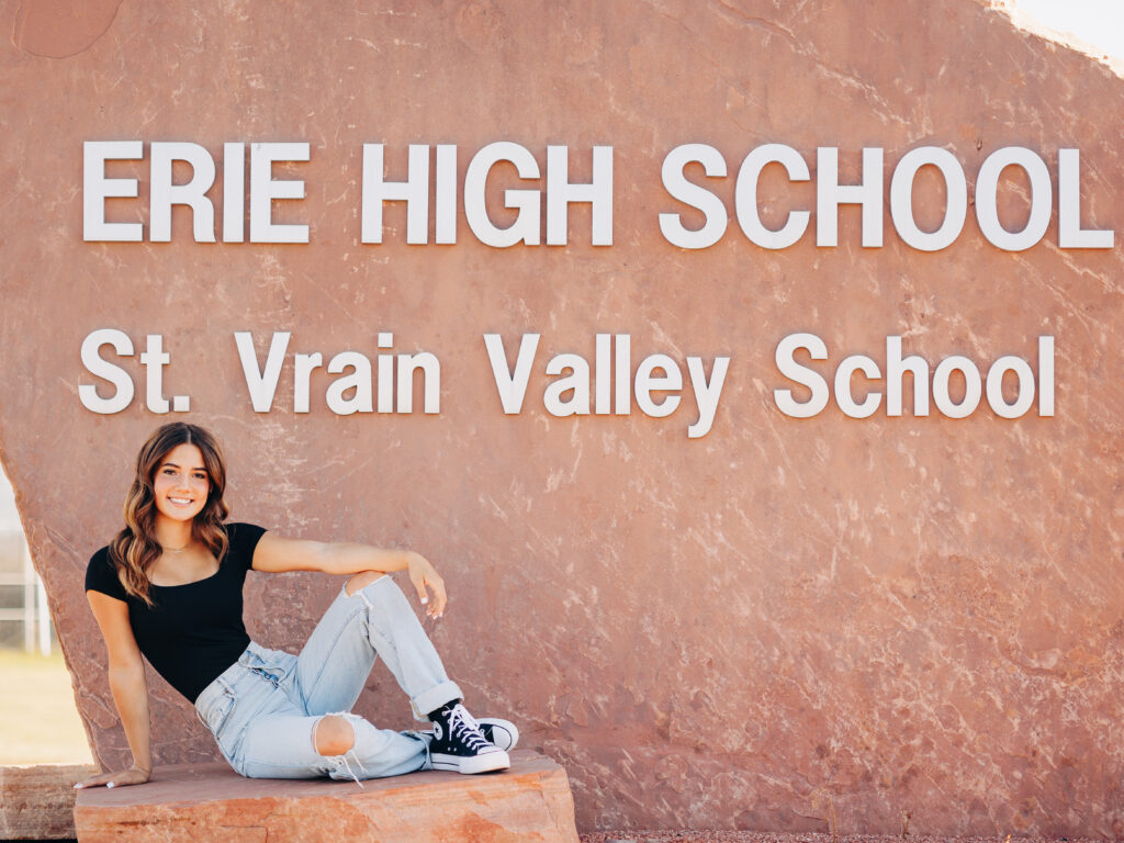 A senior wearing a black shirt and light blue jeans in Erie, CO poses at Erie High School in front of the school's sign while smiling at the camera.