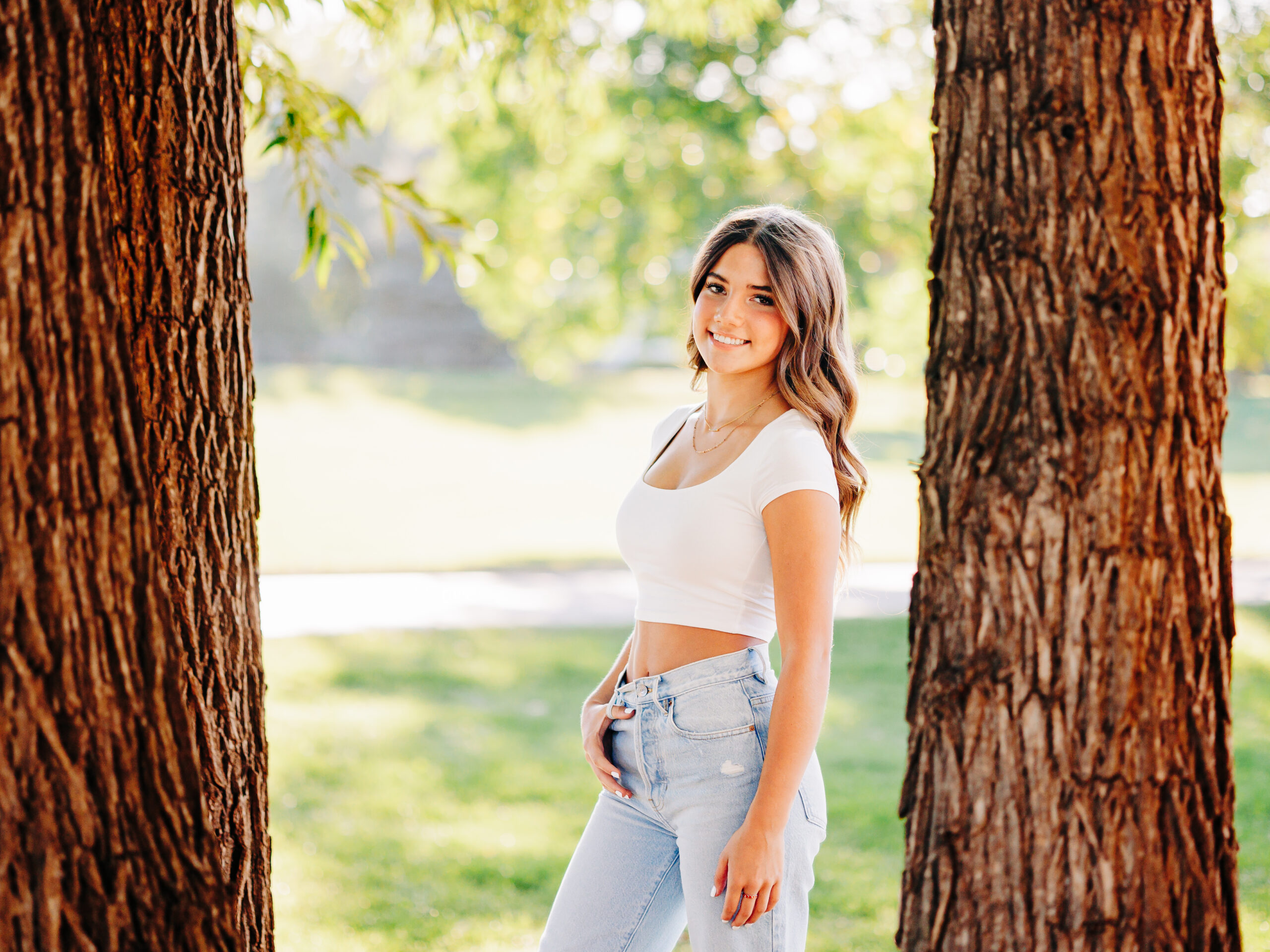 A senior wearing a white shirt and light blue jeans in Erie, CO poses at Waneka Park in between two large trees.