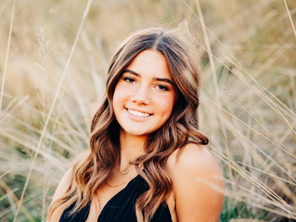 A high school senior wearing a black dress and sitting in a field of tall grass at Waneka Lake Park in Lafayette, CO.