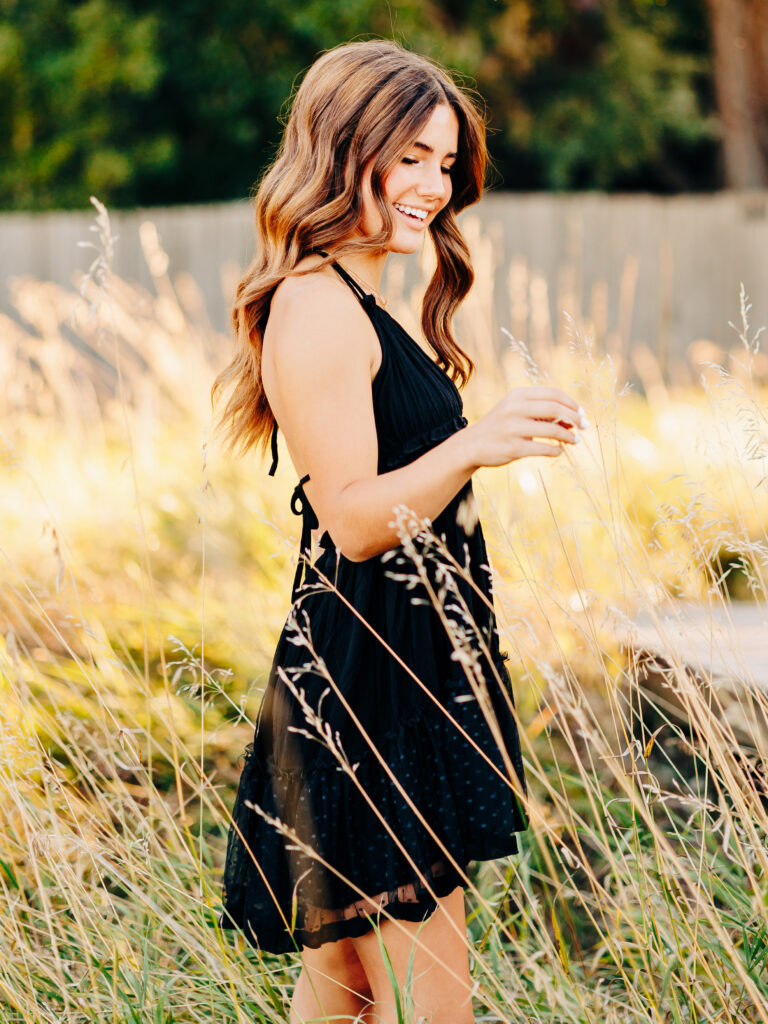 A high school senior wearing a black dress and standing in a field of tall grass at Waneka Lake Park in Lafayette, CO. She is looking down at a piece of grass in her hand.