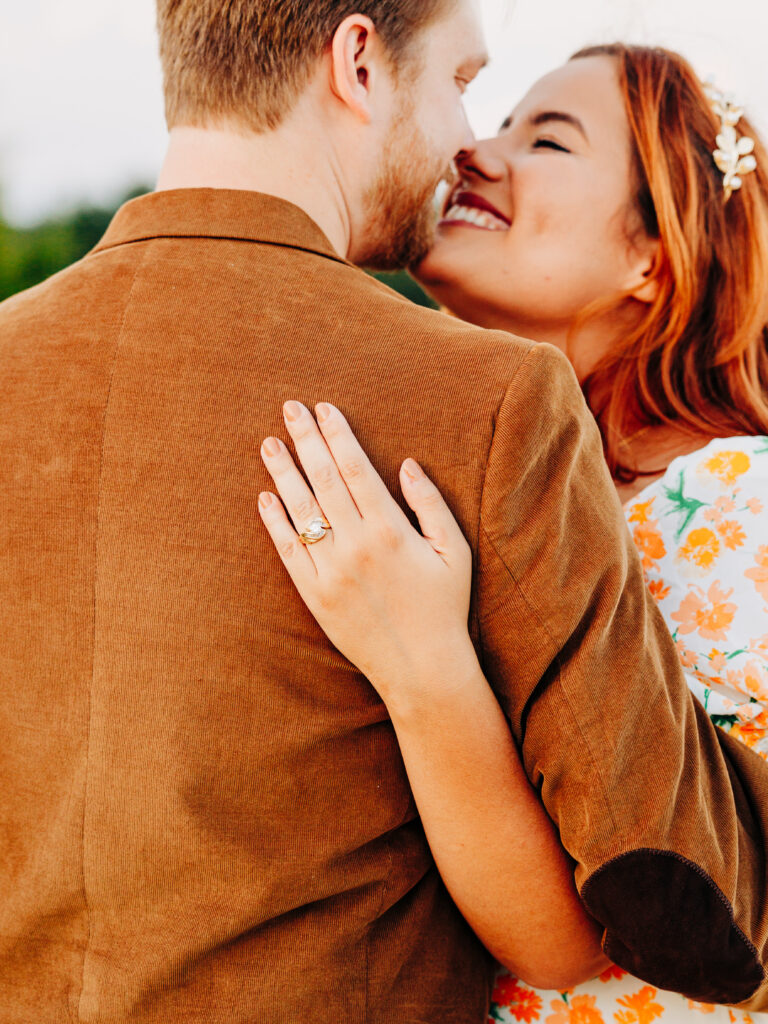 This image, taken by wedding photographer KD Captures, features an engaged couple. The man, wearing a brown jacket, is touching his nose against the woman's. The woman's hand is on the man's back, showing off her engagement ring. The image was taken at Diamond Barrel wedding venue.