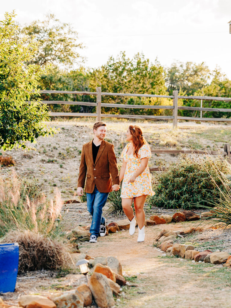 This image, taken by wedding photographer KD Captures, features an engaged couple. The man, wearing a brown jacket, holds her hand as they stroll along a path while looking at each other. The image was taken at Diamond Barrel wedding venue.