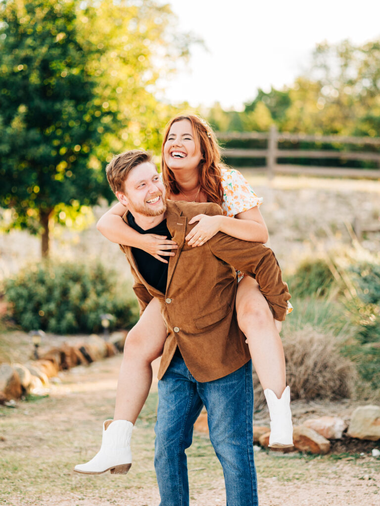 This image, taken by wedding photographer KD Captures, features an engaged couple. The man, wearing a brown jacket, is holding the woman on his back as she wraps her arms around him to hold on. She is wearing a floral dress and a floral headband. The woman is laughing. The image was taken at diamond barrel wedding venue.