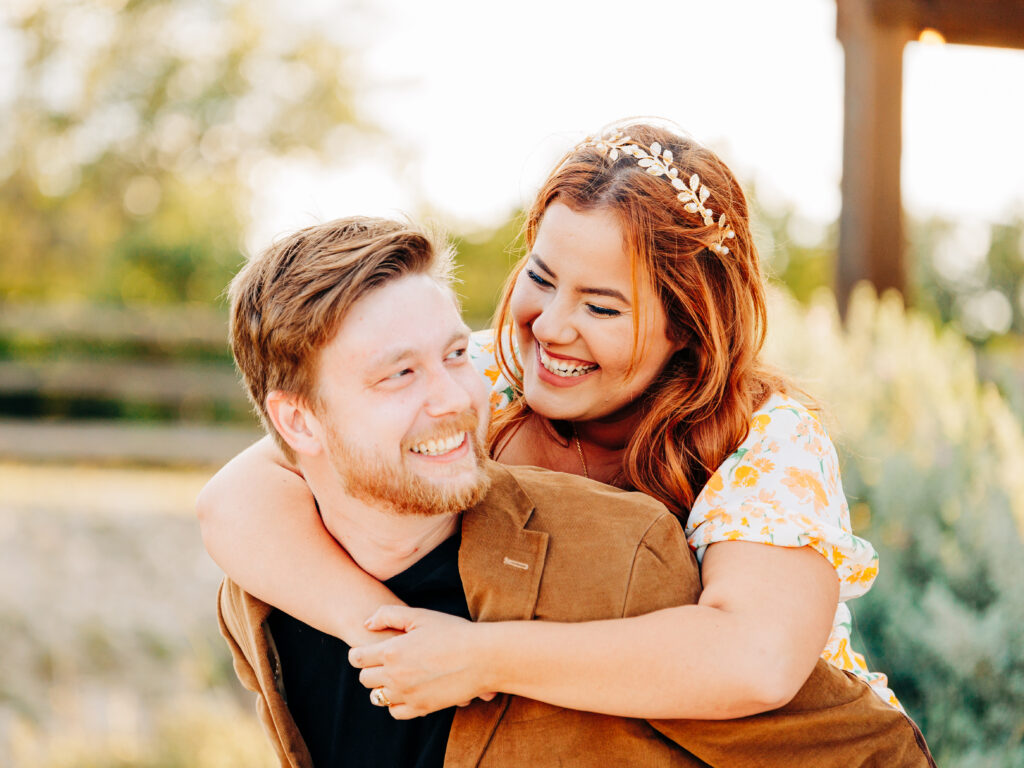This image, taken by wedding photographer KD Captures, features an engaged couple. The man, wearing a brown jacket, is holding the woman on his back as she wraps her arms around him to hold on. She is wearing a floral dress and a floral headband. The image was taken at diamond barrel wedding venue.