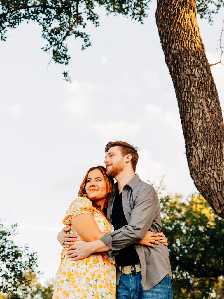This image, taken by wedding photographer KD Captures, features an engaged couple. The man is wearing a grey button up and a black tee shirt. The woman is wearing a floral dress. The couple holds each other in a tight embrace as they look off to the side. The moon is visible above them. The image was taken at Diamond Barrel wedding venue.
