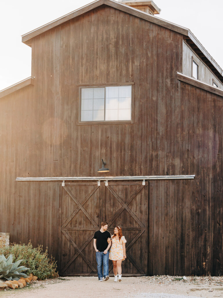 This image, taken by wedding photographer KD Captures, features an engaged couple. The man, wearing a black tee shirt, stands next to the woman while holding her hand. The woman is wearing a floral dress while they look at each other. The image was taken at Diamond Barrel wedding venue.