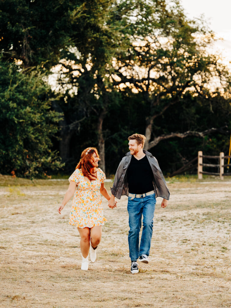 This image, taken by wedding photographer KD Captures, features an engaged couple. The man, wearing a grey button up black tee shirt, is holding the woman's hand as they run towards the camera. The woman is wearing a floral dress while they look at each other. The image was taken at Diamond Barrel wedding venue.