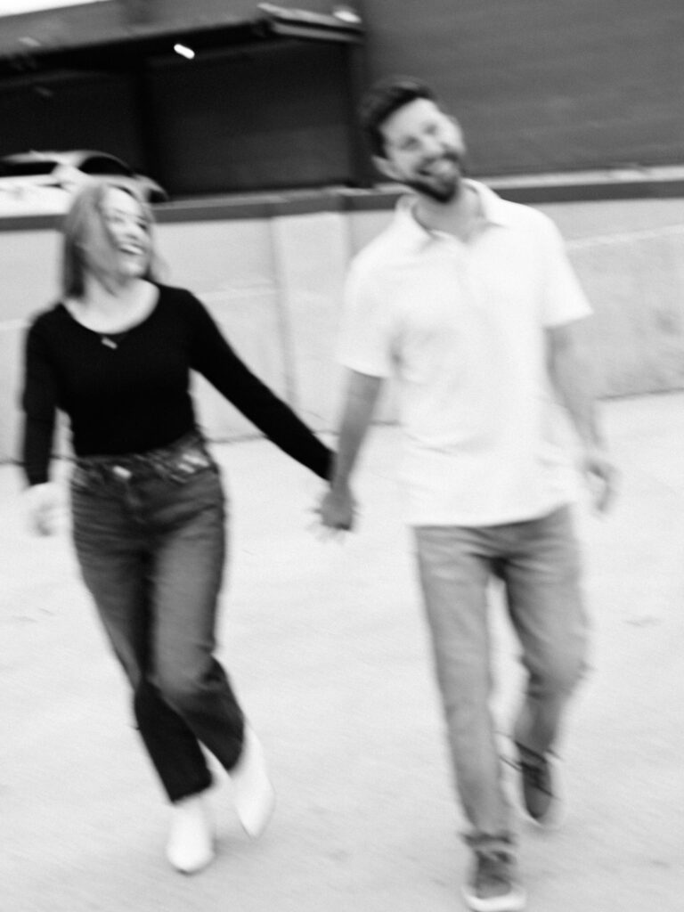 A candid, black and white motion-blurred photo of a smiling man and woman holding hands and walking. The man is dressed in a white polo shirt and jeans, while the woman wears a black long-sleeve top with jeans and white boots, both exhibiting a relaxed and joyful demeanor.