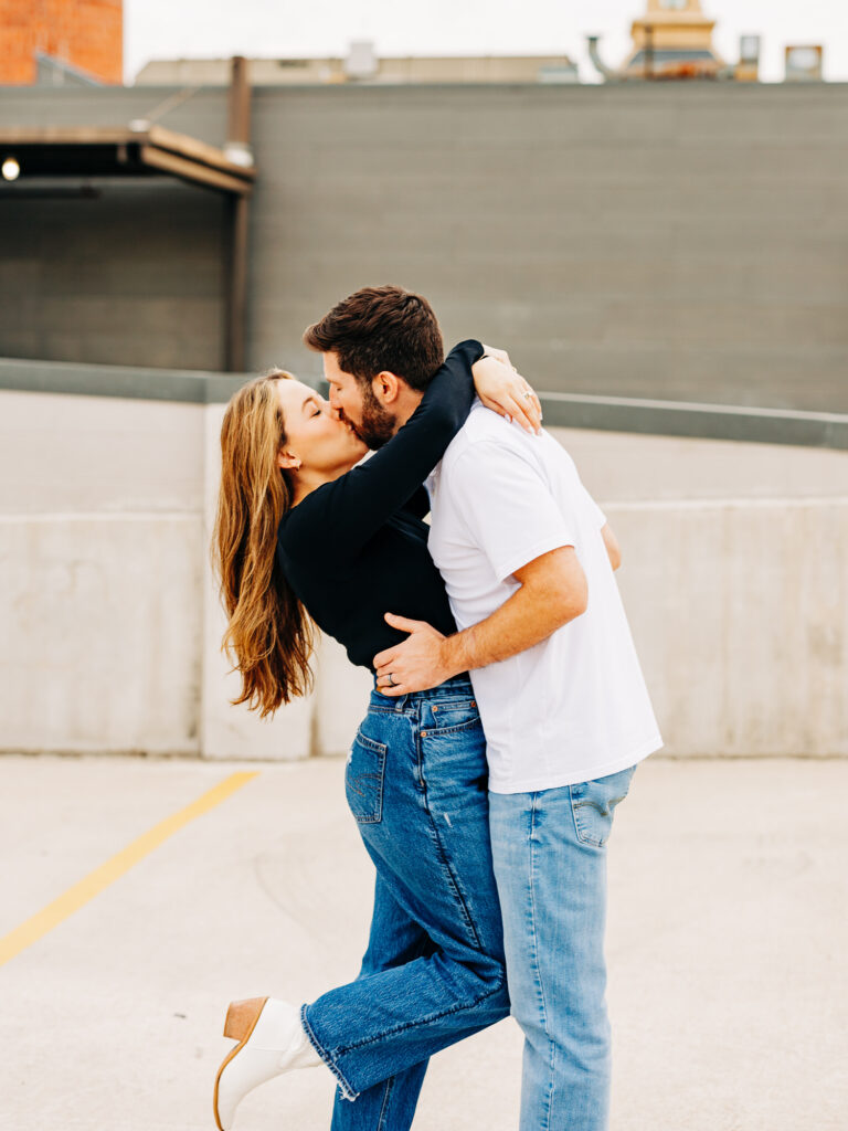 A couple stands in a parking garage in San Antonio. The couple is kissing. The woman is wrapping her arms around his neck and kicking her foot up, and the man's hands are on her waist.