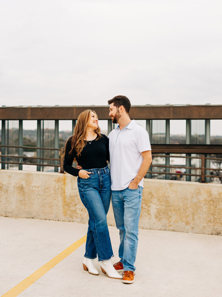 A couple stands in a parking garage in San Antonio. They are looking at each other and smiling. This is a full body image of each of them.