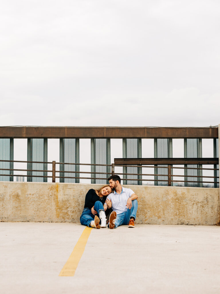 A couple sits in a parking garage and cuddles up. The woman is wrapping her arm around the man, and he is nuzzling the top of her head.