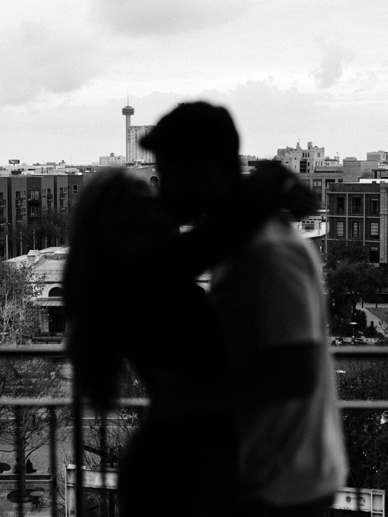 A silhouette of a couple in a close embrace is set against the urban skyline featuring the iconic Tower of the Americas. The photo, taken during a couple's session at the pearl & captured in black and white, offers a dramatic and romantic atmosphere, emphasizing the contours of the couple against the cityscape backdrop