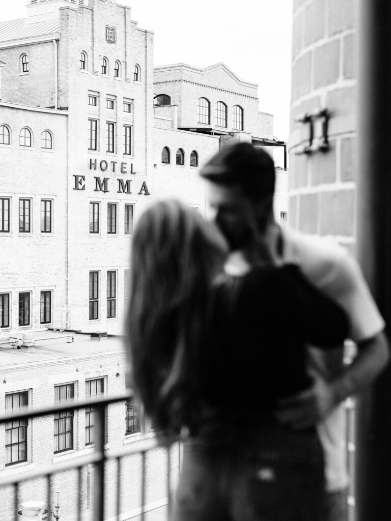 A black and white image taken during a couple's session at the pearl with a soft focus foreground of a couple in an embrace, set against the distinct backdrop of the Hotel Emma building. The couple's affectionate pose and the historic urban architecture combine to create a timeless and romantic scene
