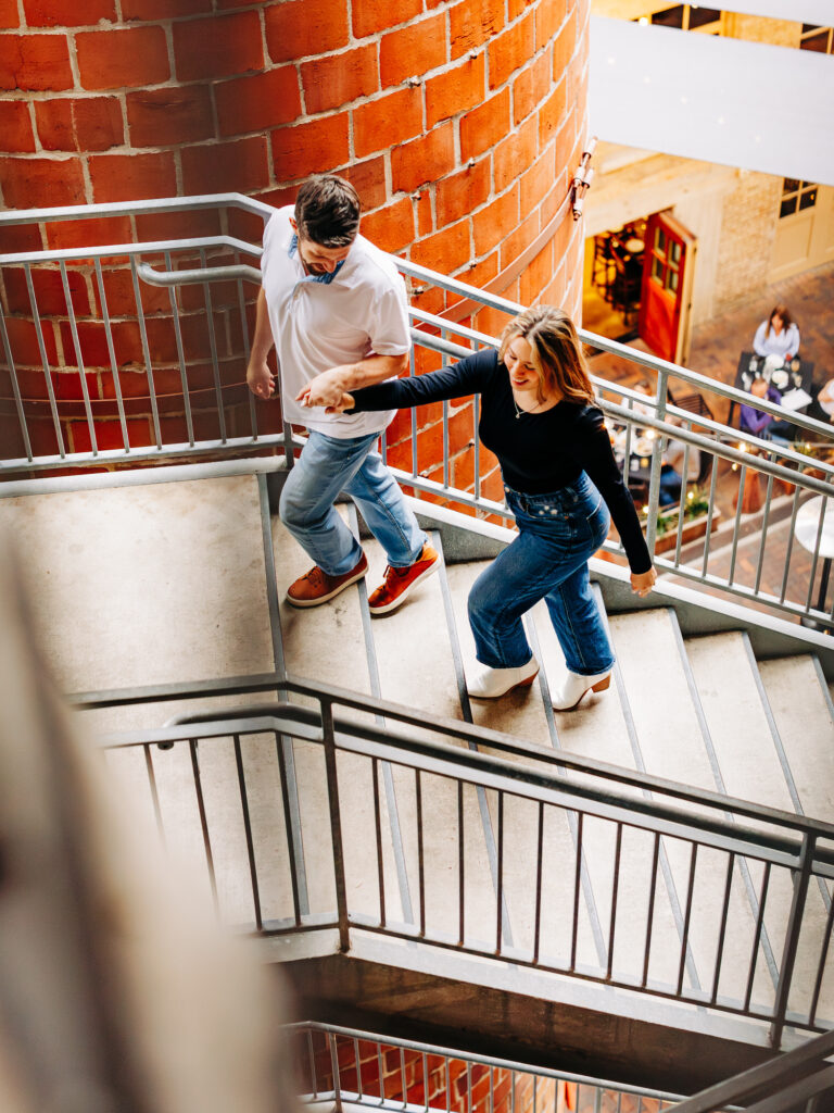 A vibrant, color photograph taken during a couple's session at the pearl from an elevated angle capturing a couple climbing a spiral staircase. The man in a white polo shirt and jeans leads the woman who is in a black top and jeans, with white boots. The warm red brick wall and the bustling cafe scene below add to the lively and cheerful atmosphere of the moment
