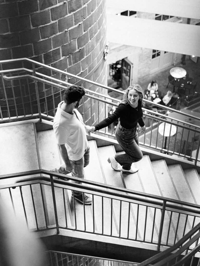 An overhead black and white image of a man and woman playfully ascending a spiral staircase. The woman, in a black top and jeans, looks back and smiles, leading the man who wears a casual white shirt and jeans. They are surrounded by the dynamic architecture of the staircase, adding a sense of movement to the scene. The image was taken during a couple's session at the pearl