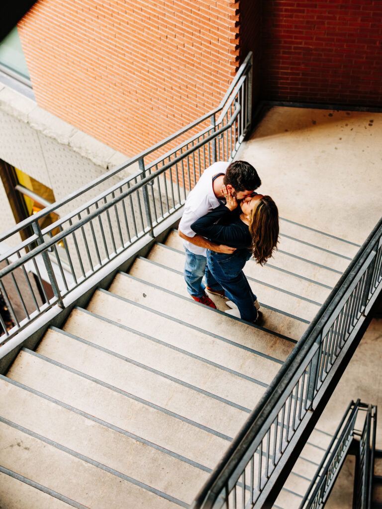 From an overhead perspective, a couple shares a romantic kiss on a stairway landing. The man is in a white polo and jeans, while the woman wears a black top and jeans. The striking contrast of the red brick wall and the geometric lines of the metal staircase frames this intimate moment beautifully. The image was taken during a couple's session at the pearl