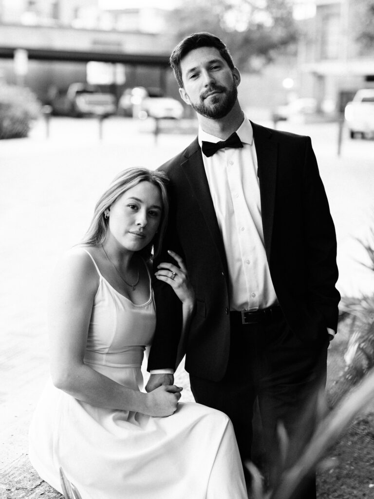 A black and white portrait of a couple taken during a couple's session at the pearl dressed in formal attire, with the woman seated and leaning against the man who stands beside her. She wears an elegant white dress, and he is in a classic black tuxedo with a bow tie. Their serene expressions and sophisticated outfits evoke a sense of timeless elegance.