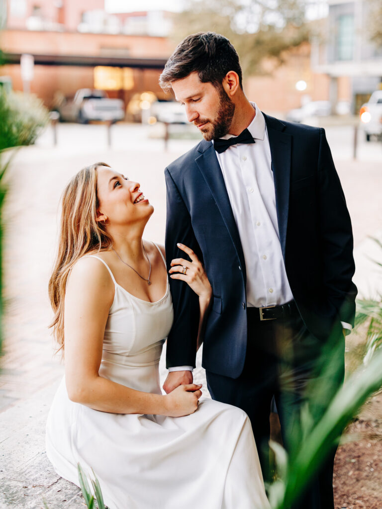 A colorful image of a woman in a white dress, seated and looking up affectionately at a man standing beside her, who is dressed in a black tuxedo and bow tie. They share a moment of connection against a softly blurred urban background, highlighting the warmth and intimacy of their relationship. The image was taken during a couple's session at the pearl