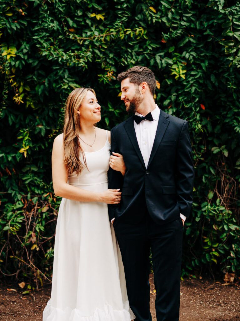 A couple stands closely together, exchanging smiles amidst a lush green backdrop of leaves. The woman, in a sleeveless white gown, gazes at the man in a sharp black tuxedo with a bow tie. Their relaxed and happy demeanors convey a sense of comfortable love and companionship.