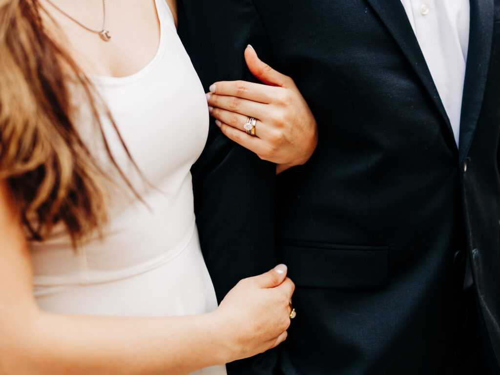 A close-up view highlighting the tender gesture of a woman's hand placed on a man's black suit jacket. The woman's white dress, elegant necklace, and engagement ring are visible, suggesting a special occasion. The image conveys closeness and the fine details of a couple's attire on a celebratory day