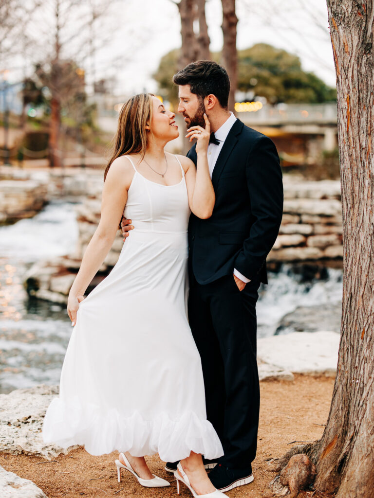 An affectionate moment captured between a couple by a riverside setting, with the woman in a white dress and heels leaning towards the man in a black suit and bow tie. They stand close to each other, sharing a gaze that speaks of affection and intimacy, with the natural beauty of the riverbank and trees softly blurred in the background.