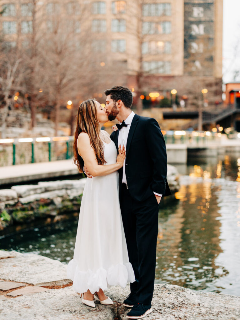 A bride and groom stand in front of the Riverwalk kissing. The woman is resting her hand on his chest