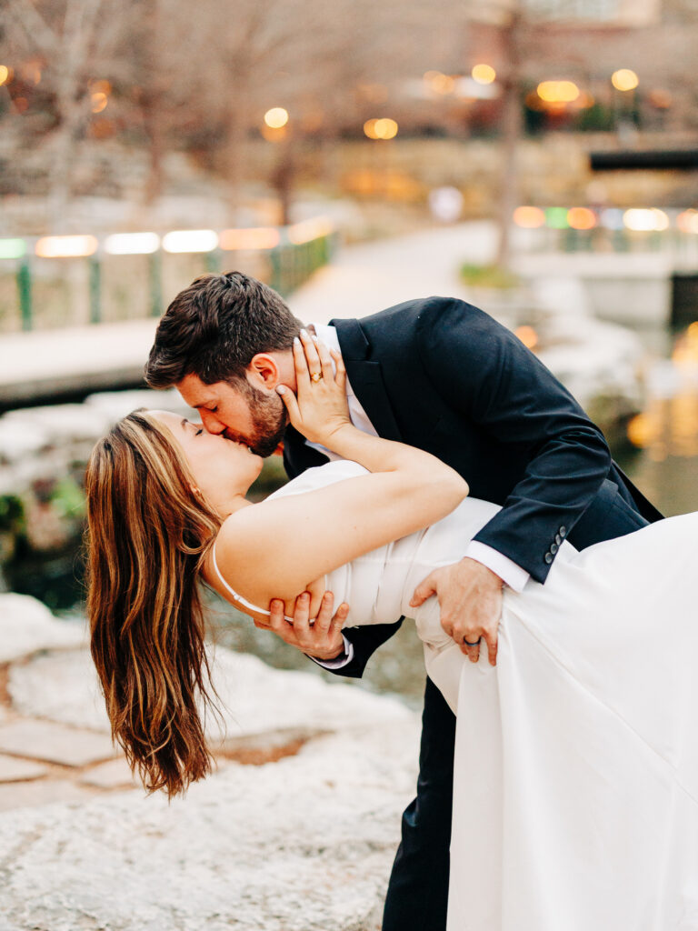 A bride and groom stand in front of the Riverwalk kissing. The man is dipping the woman and she has her hand on his face.