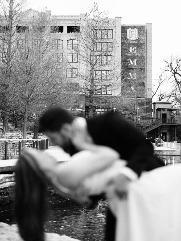 A bride and groom stand in front of the Riverwalk kissing. The man is dipping the woman and she has her hand on his face. The couple is out of focus and Hotel Emma is in focus in the background. The image is in Black and white.