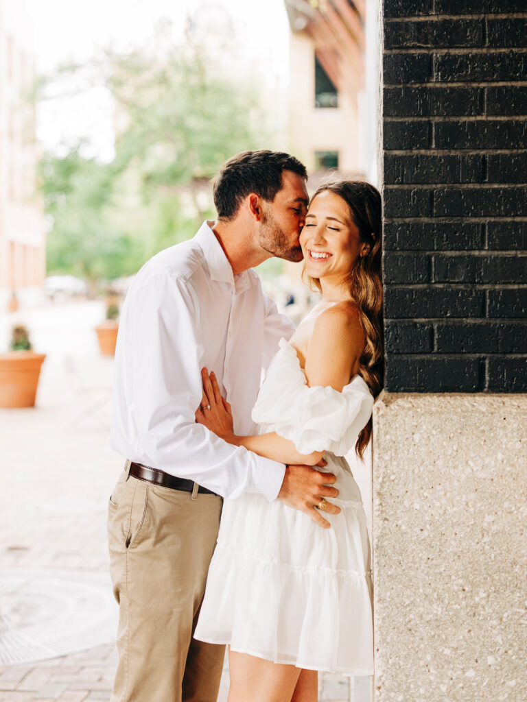 A photo of an engaged couple in San Antonio. The woman has her back against a brick wall, and has her hand positioned on the man's stomach. The man is kissing the woman's cheek as she smiles.
