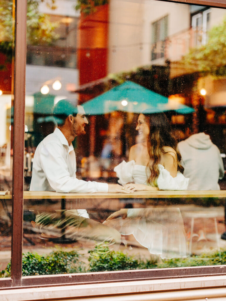 A photo of an engaged couple sitting inside of a coffee shop at a table together. The photo was taken through a window from the outside of the shop. The man and woman are looking at eachother and holding hands.