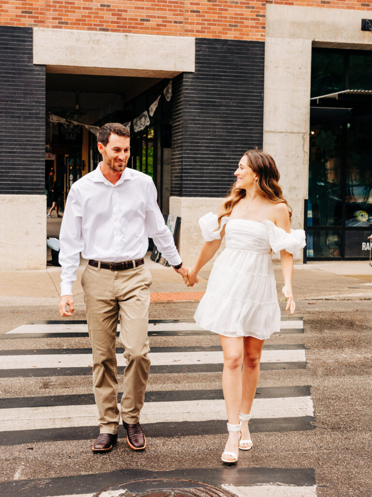 A photo of a man and a woman walking across a crosswalk in downtown San Antonio. The woman is wearing a white dress and white heels and the man is wearing a white button-up and khaki pants.
