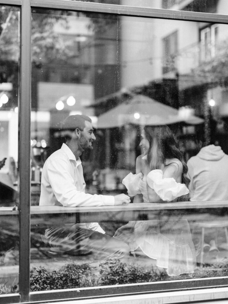 A photo of an engaged couple sitting inside of a coffee shop at a table together. The photo was taken through a window from the outside of the shop. The man and woman are looking at eachother lovingly. The photo is monochrome.