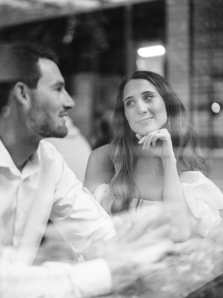 A monochrome photo of an engaged couple sitting together inside of a coffee shop. The woman is resting her chin on her hand as she lovingly looks at the man, who is telling a story. The photo was taken through a window from the outside of the coffee shop.