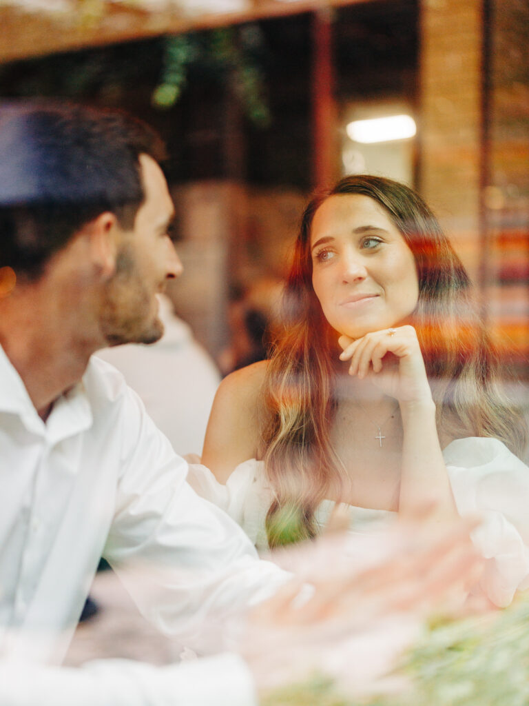 A photo of an engaged couple sitting together inside of a coffee shop. The woman is resting her chin on her hand as she lovingly looks at the man, who is telling a story. The photo was taken through a window from the outside of the coffee shop.