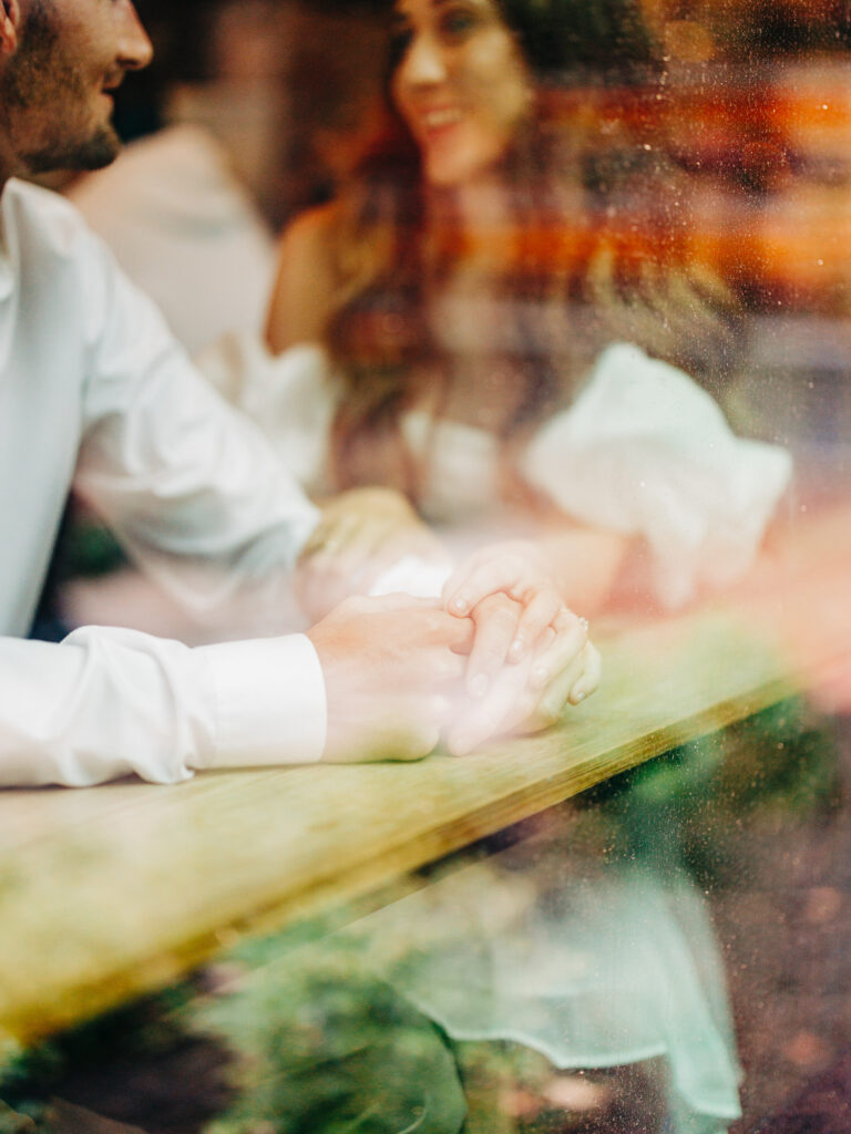 A photo of an engaged couple sitting together inside of a coffee shop. The image is focused on their hands, as they hold each other's. The photo was taken through a window from the outside of the coffee shop.