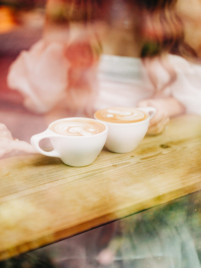 A photo of an engaged couple sitting together inside of a coffee shop. The image is focused on their lattes as the couple talks. The photo was taken through a window from the outside of the coffee shop.