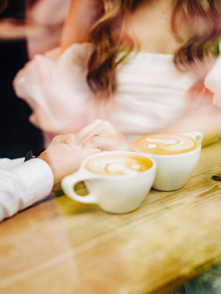 A photo of an engaged couple sitting together inside of a coffee shop. The image is focused on their lattes as the couple talks and holds hands. The photo was taken through a window from the outside of the coffee shop.