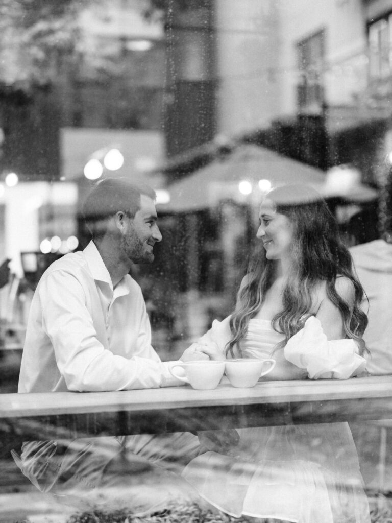 A photo of an engaged couple sitting together inside of a coffee shop. The image shows the couple with two coffees as they hold hands and lovingly look at each other. The photo was taken through a window from the outside of the coffee shop.