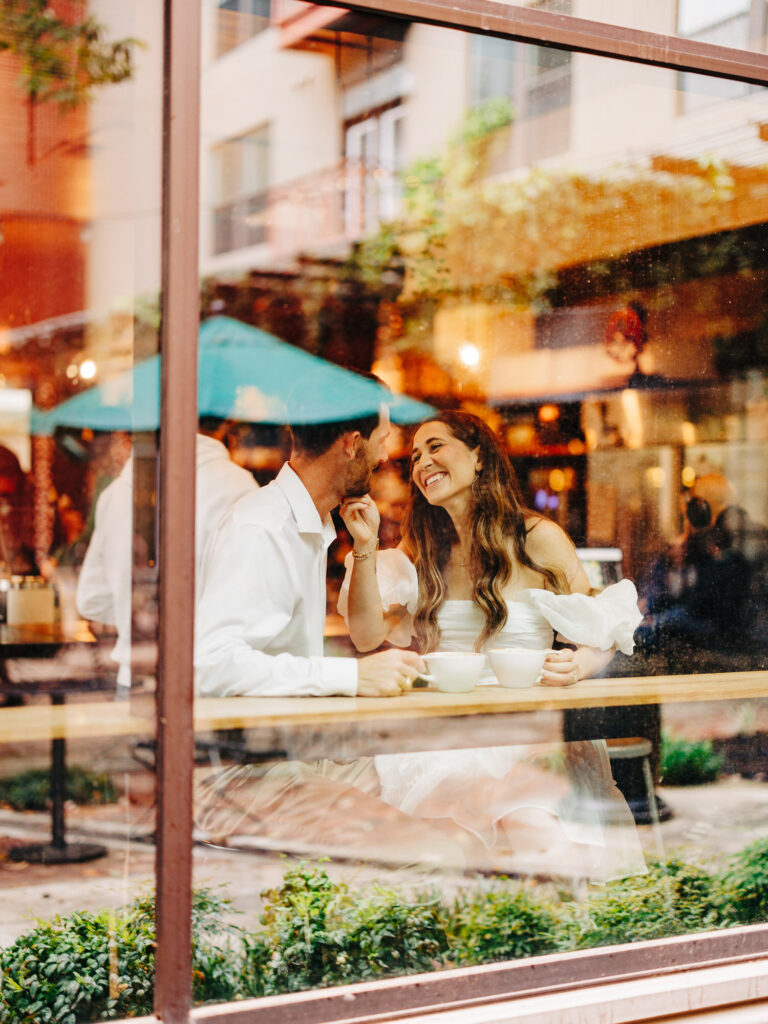 A photo of an engaged couple sitting inside of a coffee shop at a table together. The photo was taken through a window from the outside of the shop. The man and woman are looking at eachother, and the woman is holding his chin while they both hold the handles of their cups.