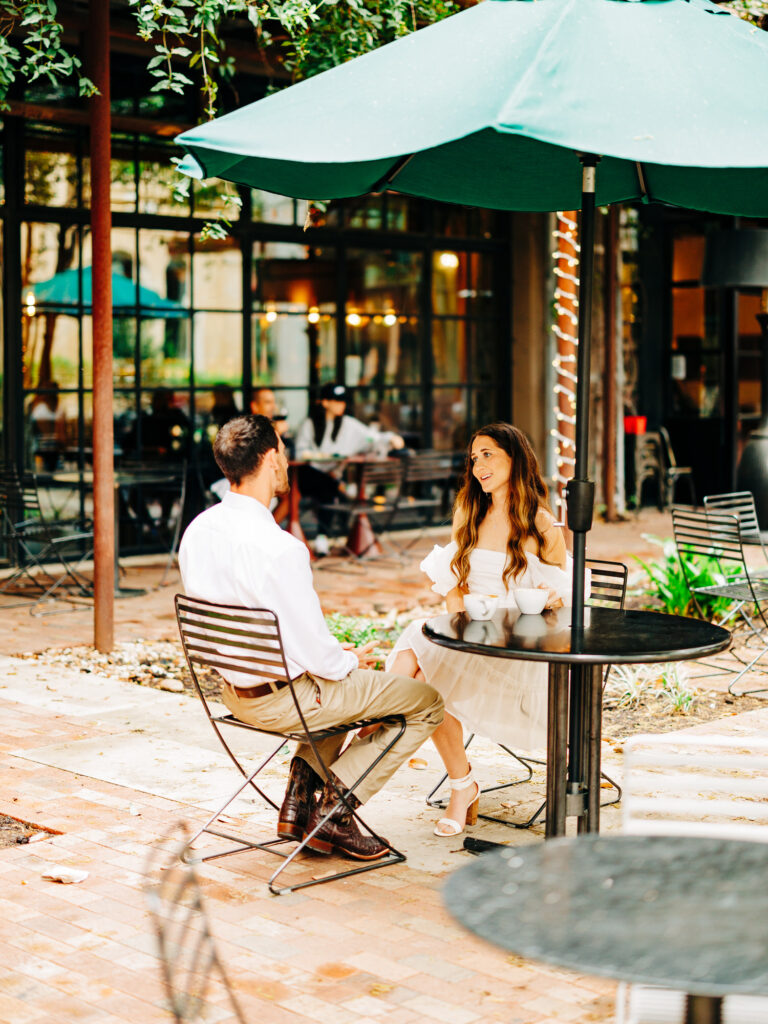 A man and a woman sitting at a patio table outside of a coffee shop. They are looking at eachother and engaging in conversation.