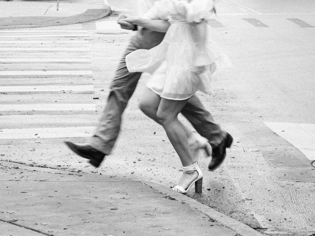 A monochrome motion-blur image of a couple jumping up onto a curb after crossing the street. The photo only shows them from the waist down.