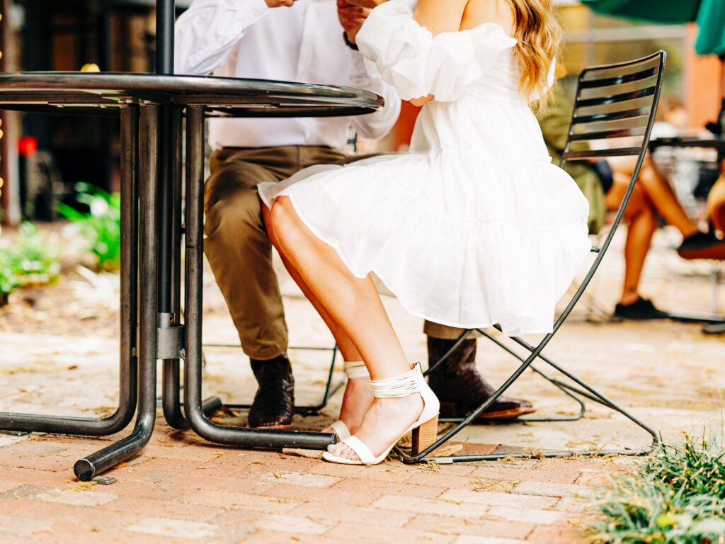 A man and a woman sitting at a patio table outside of a coffee shop. They are looking at eachother and engaging in conversation. The photo is focused on their feet, from the waist down.