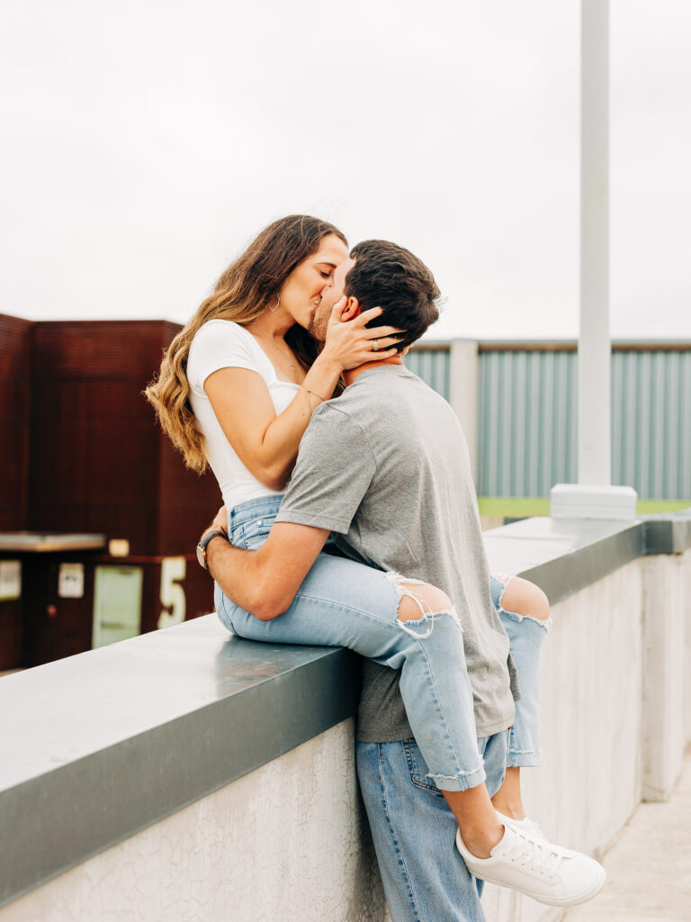 A man and a woman in a rooftop parking garage in San Antonio. The man is wearing a grey t-shirt and jeans, and the woman is wearing a white shirt and ripped jeans. The woman is sitting on a railing and wrapping her legs around the man, while holding onto his face as they kiss. The man is holding onto her waist.